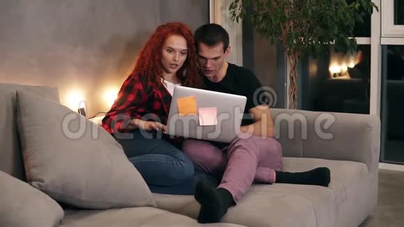 漂亮的年轻夫妇坐在一起沙发上有一台灰色的笔记本电脑一对短发男人和红发女孩在看视频的预览图