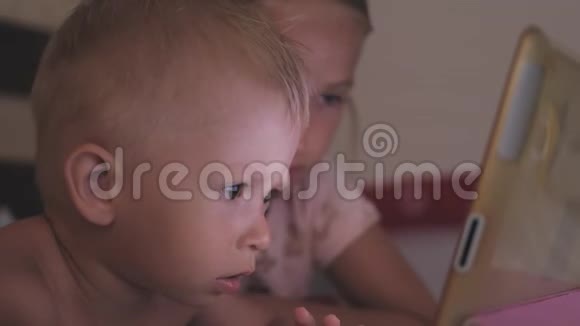两个孩子躺在床上在黑暗的光线下在社交网络上玩平板电脑把小男孩和他姐姐关起来视频的预览图
