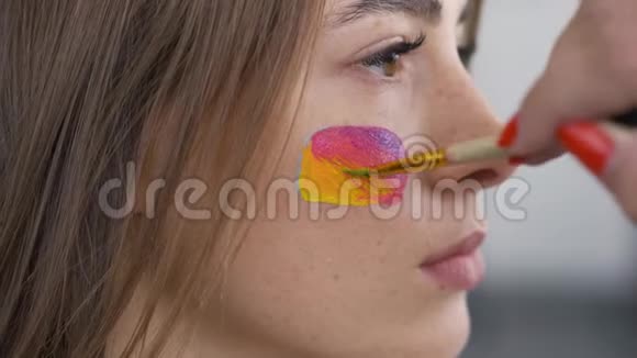 一个年轻漂亮女孩的详细肖像艺术家用小软笔在女孩脸上画多色图标视频的预览图