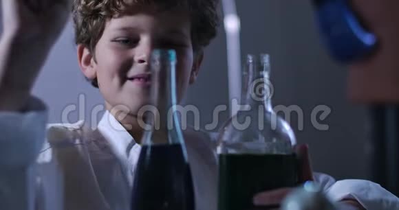年轻白种人化学家混合化学品的特写头发卷曲的男孩穿着实验室外套手里拿着装满液体的瓶子视频的预览图