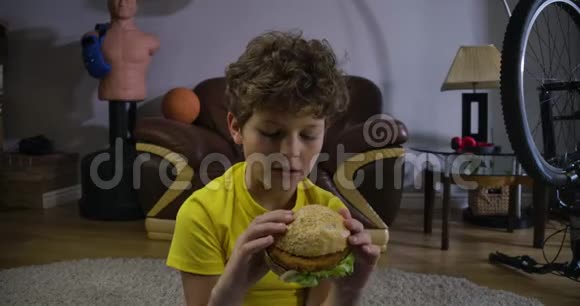 白人男孩在电视前咬汉堡包的特写镜头十几岁的卷发小孩在家看电影视频的预览图