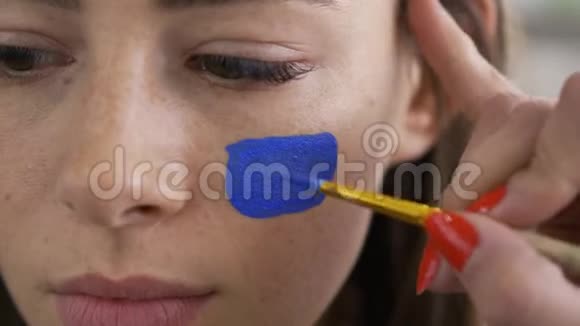 一个年轻漂亮女人的死脸艺术家用小软刷在女孩脸上画蓝色图标身体艺术视频的预览图