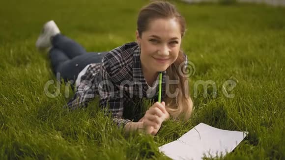 漂亮漂亮的女孩躺在草地上在笔记本上记笔记咀嚼小玩意看着相机微笑4K视频的预览图