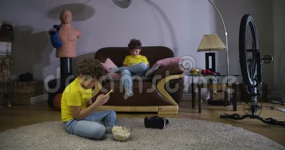使用智能手机穿休闲服装的十几岁白种人男孩的双重暴露儿童坐在沙发和地毯上视频的预览图