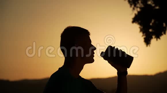 夕阳下一个健康的年轻人背着太阳喝水的剪影缓缓移动视频的预览图
