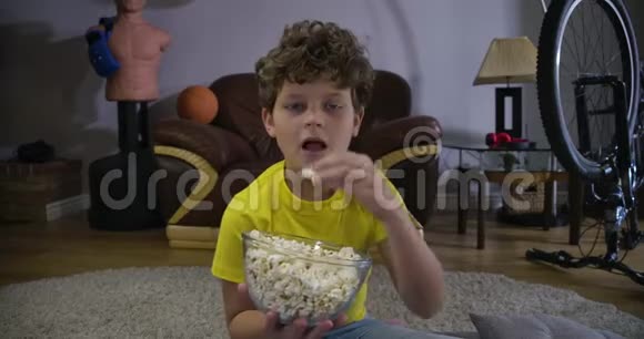 小白种人男孩看电视和嚼爆米花的肖像可爱的卷发少年在家吃垃圾食品视频的预览图