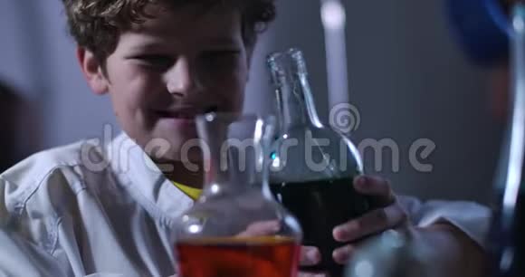 白种人男孩混合黑色和红色化学试剂的特写脸穿着实验室外套拿着装满瓶子的少年视频的预览图