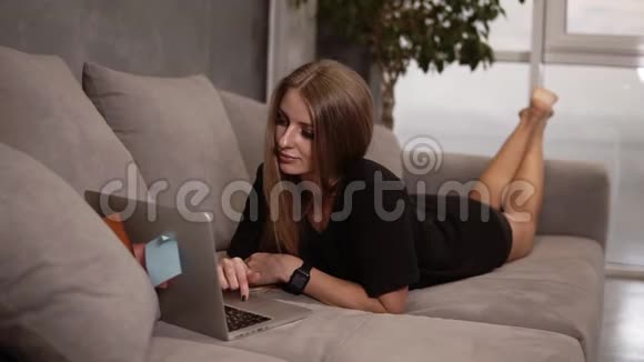 漂亮的长发快乐的白种人女士躺在舒适的现代灰色沙发上在上面看有趣的照片视频的预览图