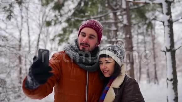 年轻可爱的时髦夫妇在森林里的大雪下为社交媒体做现场直播带智能手机的博客男视频的预览图