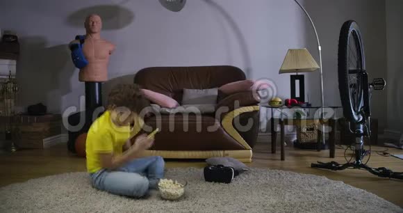 白人青少年使用智能手机和吃爆米花的时间推移一个卷发的小男孩坐在地板上视频的预览图