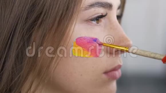一个年轻漂亮女人的详细肖像艺术家用小软笔在女孩脸上画多色图标视频的预览图