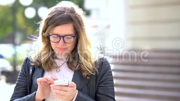 一个漂亮的女孩戴着眼镜站着在智能手机的帮助下交流微笑好消息视频的预览图
