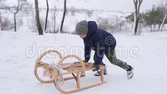 穿着温暖衣服的男孩在下雪的冬天把老式木雪橇推到山上小男孩在公园里雪橇视频的预览图