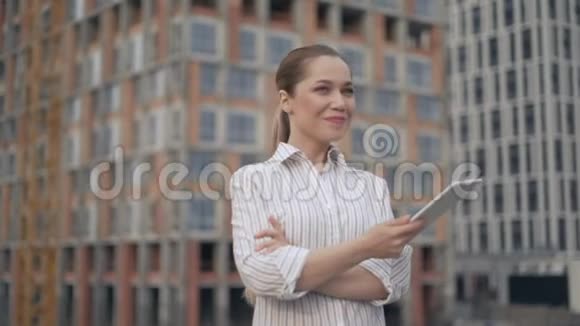 一个自信快乐的女人在一栋正在建造的多层建筑的背景下一个女人拿着一块平板电脑在她身上视频的预览图