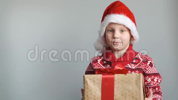 戴着圣诞帽的可爱男孩戴着灰色背景的礼盒看着相机微笑着视频的预览图