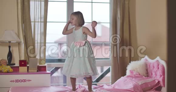 快乐的小白种人女孩穿着优雅的裙子在家里的粉红色床上跳跃和旋转玩耍的孩子周末视频的预览图