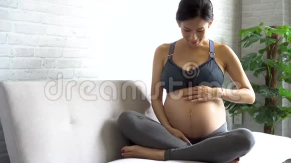 年轻的亚洲孕妇期待着新的孩子坐在家里的沙发上视频的预览图