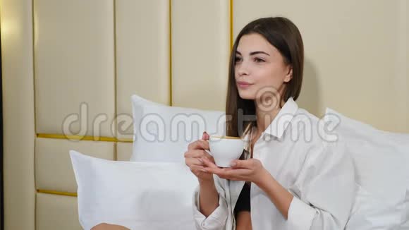 中等镜头的快乐微笑的女人享受华丽的早晨在卧室床上喝茶或咖啡穿白色衣服视频的预览图