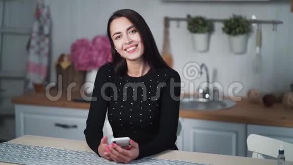 坐在厨房里的微笑女人看着摄像机手里拿着电话视频的预览图
