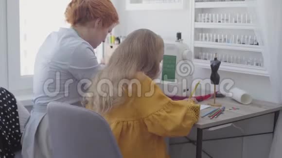 可爱的小女孩坐在前台的桌子旁红发的妈妈在旁边缝衣服视频的预览图