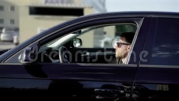 一个留着胡子的年轻人坐在他的黑色汽车上的停车场里商用轿车静静地沿着其他机器行驶视频的预览图