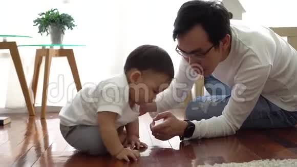 严肃的亚洲小男孩在地板上爬行而年轻的父亲坐在他旁边的客厅里视频的预览图