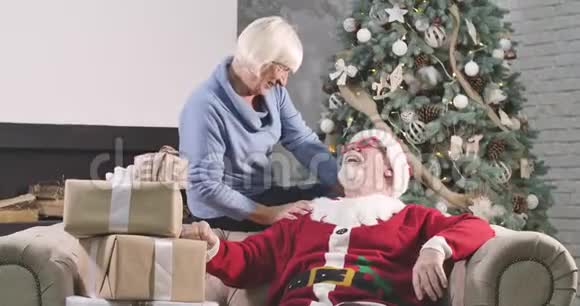 戴着红色圣诞帽的高白种人快乐金发女人坐在沙发上手里拿着礼品盒看着视频的预览图