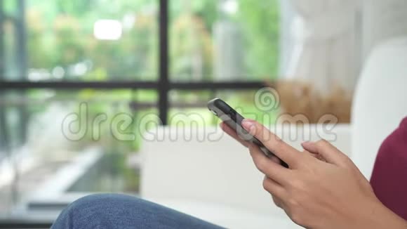 美丽的亚洲女人享受时光坐在窗前的现代沙发上用智能手机在客厅放松视频的预览图