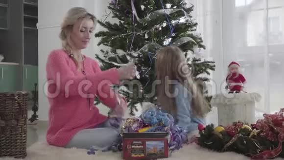 白种人金发女人和黑发女孩坐在新年树前的地板上选择花环母亲和视频的预览图