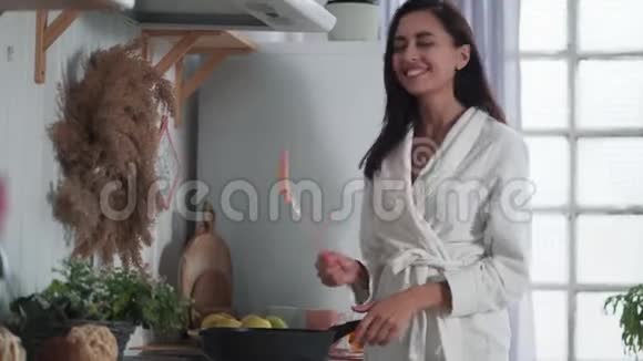穿着白色浴袍在厨房做饭在家跳舞的年轻女子视频的预览图