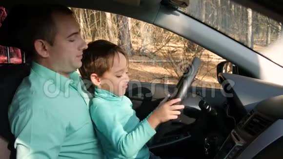 一个可爱的男孩转动汽车的轮子坐在他父亲腿上父亲教儿子汽车的装置视频的预览图