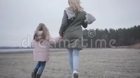 后景的成年白种人妇女和小女孩穿着温暖的衣服沿着秋天的海滩奔跑高加索人视频的预览图