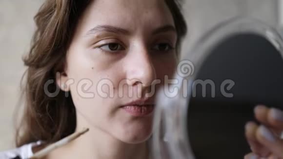 一个年轻的女人在她的眼睛下面用刷子涂抹遮瑕膏然后看着一面小镜子基本化妆视频的预览图