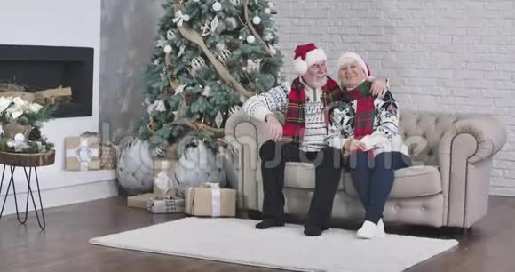 一对快乐的白人夫妇坐在圣诞树旁边的沙发上看着摄像机和礼物视频的预览图