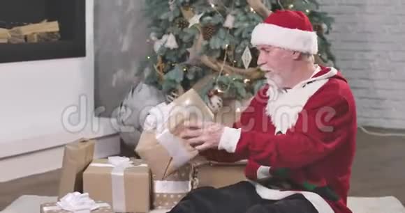 白种人留着白胡子和胡子拿着礼品盒摇着它戴着红帽子和圣诞毛衣的圣诞老人视频的预览图