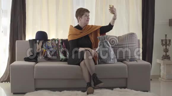 成年白人女性通过智能手机与用户交谈漂亮的女士坐在沙发上带着礼品袋和视频的预览图