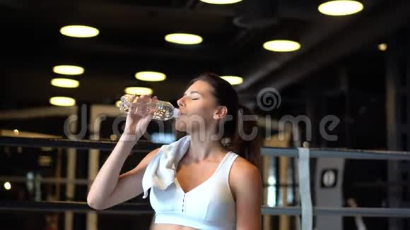 漂亮的年轻女人肩膀上有一条毛巾在健身房里喝着瓶子里的水视频的预览图