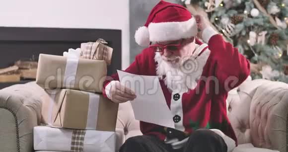 聚焦圣诞老人坐在沙发前的圣诞树和阅读信件戴红眼镜的成熟男人视频的预览图