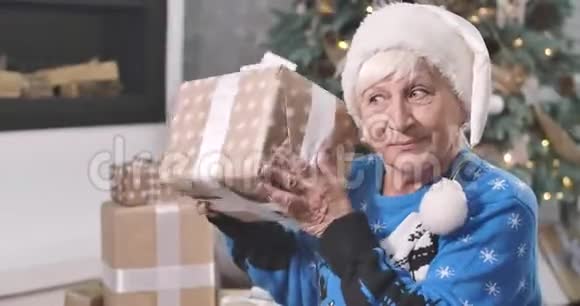 好奇的白人老妇人戴着圣诞帽摇着礼盒微笑着的肖像快乐的女士坐在前面视频的预览图