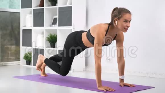 赤脚瑜伽女士戴着耳机在白色家庭内部的垫子上做运动视频的预览图