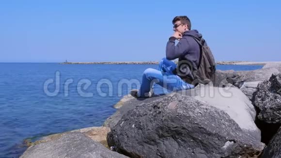 海滨的男人拿着带三脚架的背包手机拍摄自拍视频视频的预览图