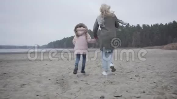 镜头跟随成年女子和穿着温暖衣服的小女孩在秋天的海滩上奔跑快乐的白人母亲和视频的预览图