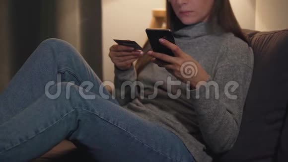 女人躺在舒适的房间里的沙发上用信用卡和智能手机在线购买网上在线视频的预览图
