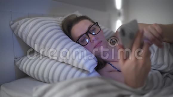 戴眼镜的女人躺在床上用智能手机她揉眼睛因为她又累又困概念视频的预览图