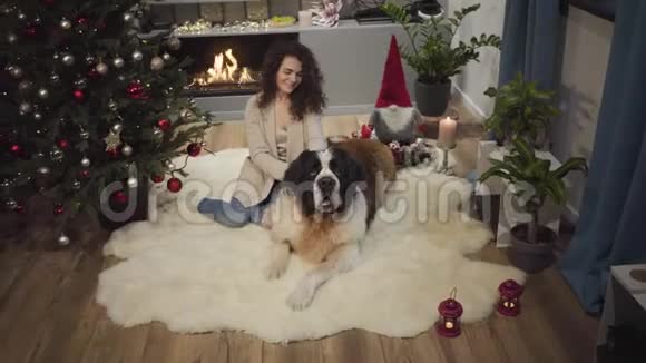 笑容满面的白种人女孩和大狗坐在壁炉前的软地毯上装饰圣诞树视频的预览图