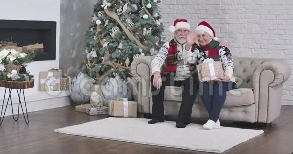 中长镜头的快乐圣诞老人和夫人坐在沙发上拿着礼物对着镜头挥手快乐的老人视频的预览图