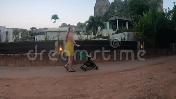 侧视一位穿着黄色衣服的年轻母亲带着婴儿车在里面散步在海岸的木板路上视频的预览图