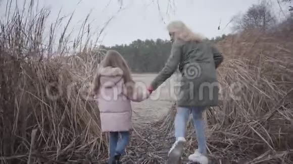 镜头跟随成年白种人妇女和女孩通过灌木丛走到湖边或河流漂亮的妈妈和可爱的视频的预览图