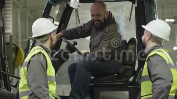 两名戴着头盔和背心的白种人男性工人坐在牵引车上与留胡子的同事交谈工厂雇员视频的预览图