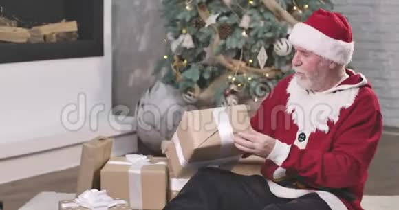 圣诞老人坐在圣诞树的背景和摇动的礼盒的肖像戴红帽子的好奇老人视频的预览图